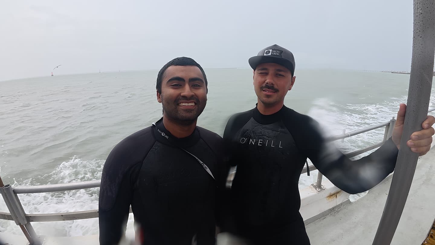 Two wetsuit-clad SPI Dive Services crew members smile together on a boat in choppy coastal water.