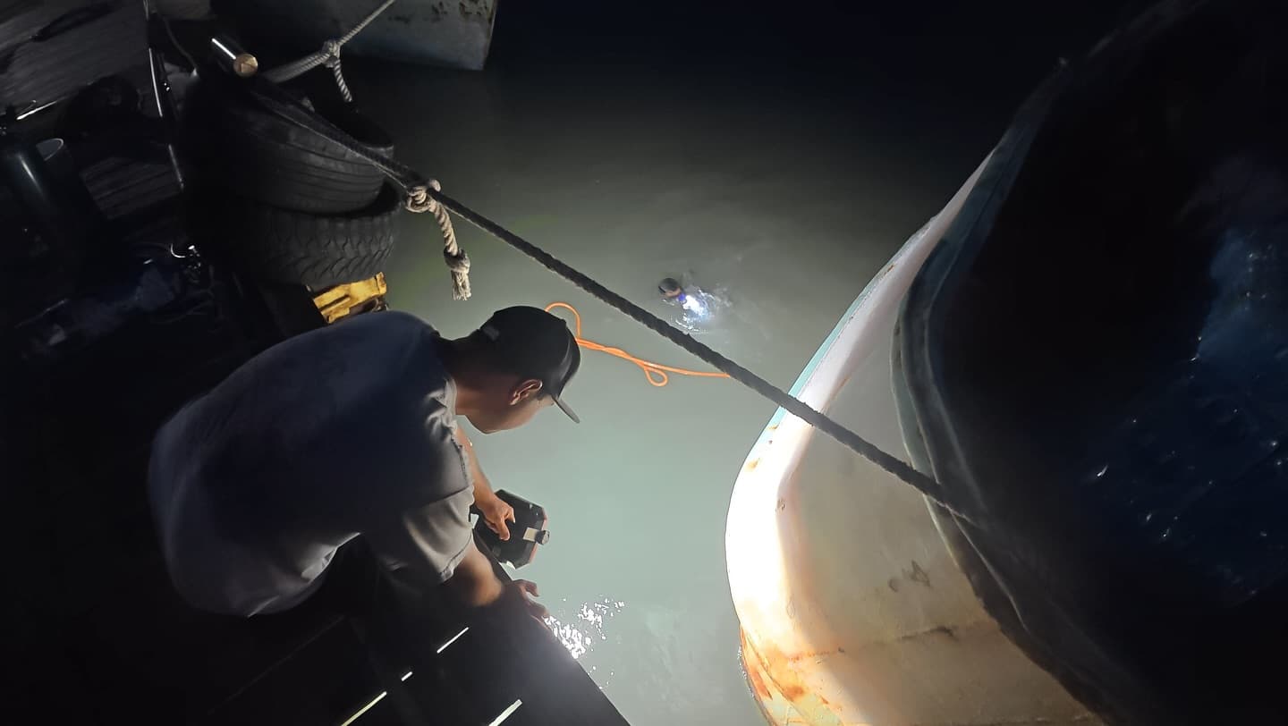 Dockside crew member watches a diver work by headlamp in dark water beside a boat at night.