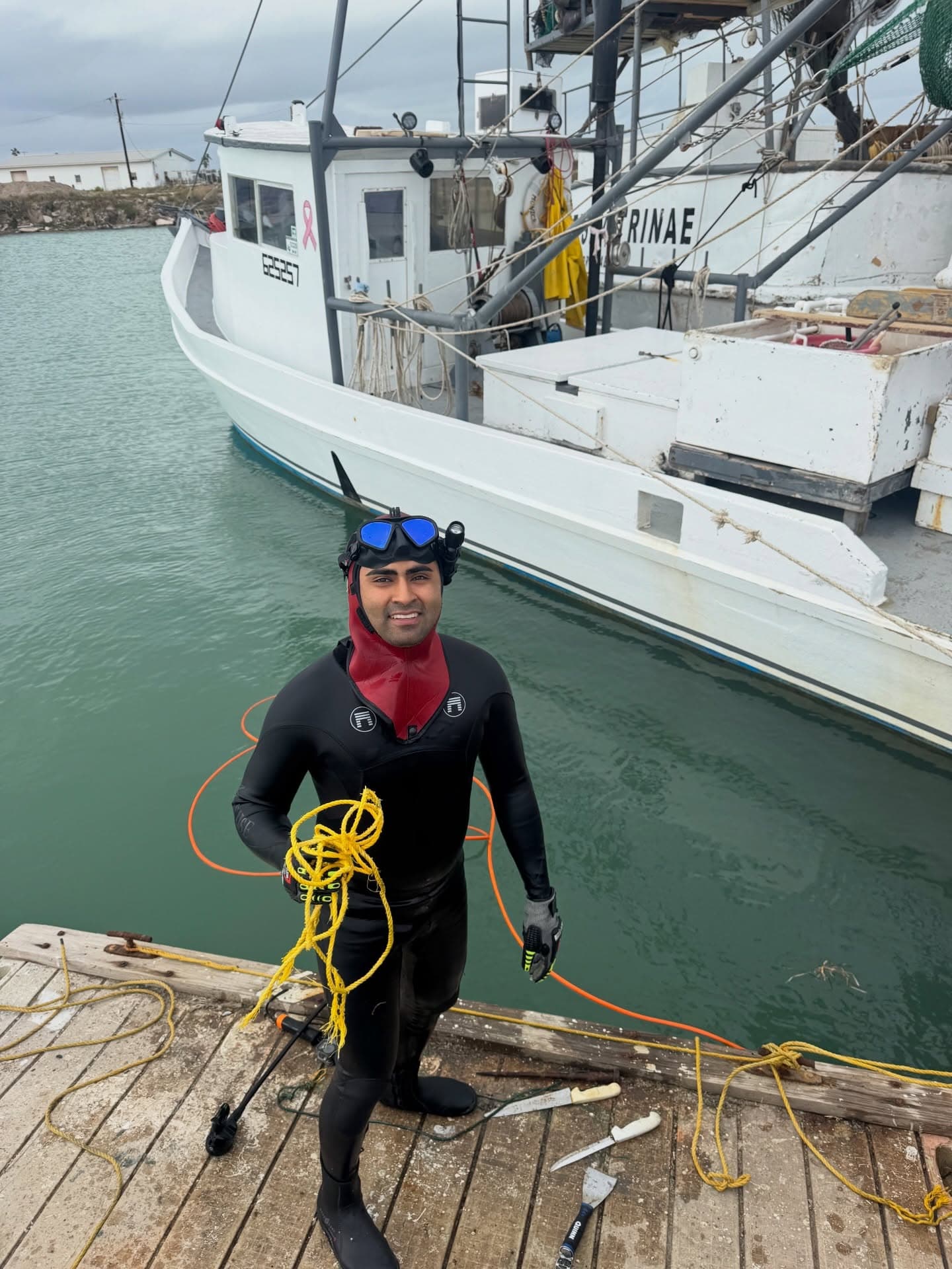SPI Dive Services diver in a black wetsuit stands ready on a dock beside a commercial fishing boat.