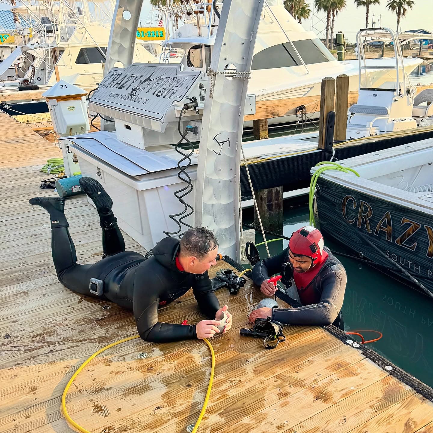 Two divers prepare equipment on a wet marina dock beside the sportfishing boat Crazy Fish.