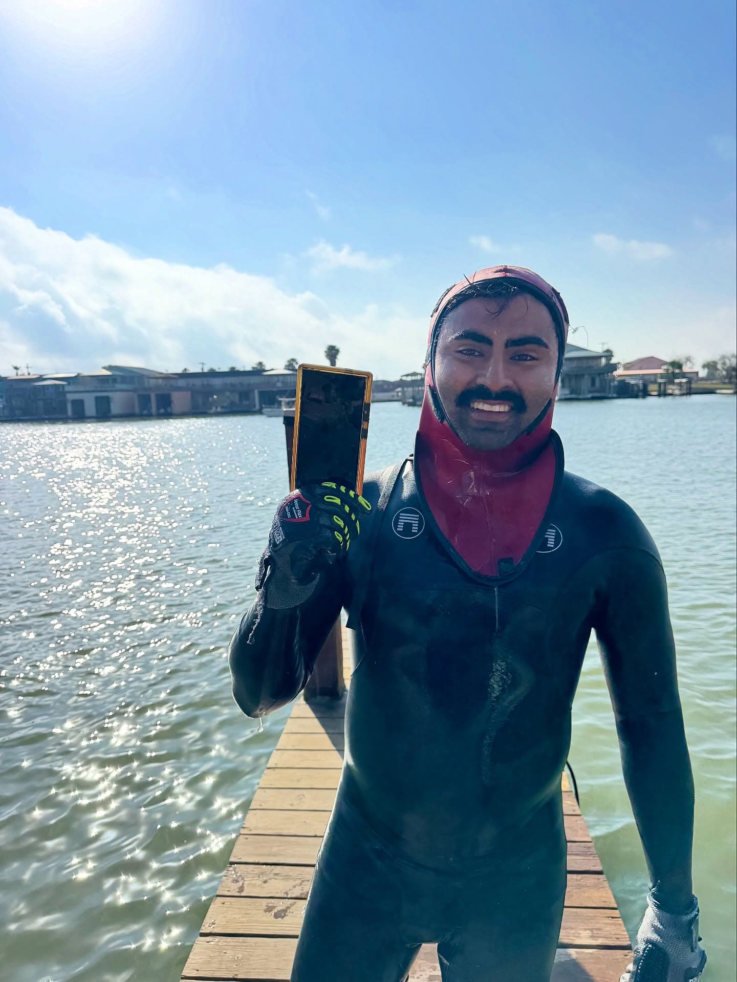 Smiling diver in a black wetsuit and red hood holds up a removed rectangular marine anode on a sunny dock.