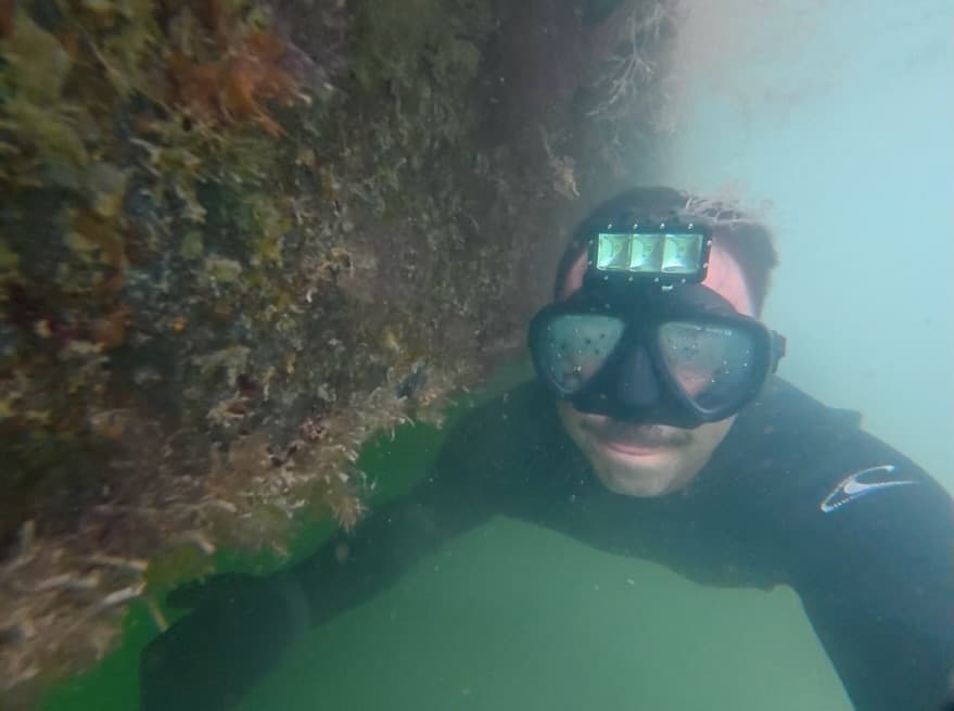 SPI Dive Services diver wearing a mask and headlamp works underwater beside a marine-growth-covered hull.